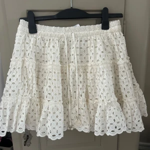Vit broderad kjol med volang - Supersöt vit kjol med broderade detaljer och volangkant. Kjolen har resår i midjan och är kort i modellen, perfekt för en fräsch och somrig look. Lätt och luftig med fina hålmönster och romantisk känsla.