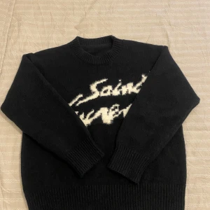 Svart stickad tröja från Saint Laurent - Svart stickad tröja från Saint Laurent med vit, suddig logotyp framtill. Klassisk rund halsringning och långa ärmar med ribbade muddar och nederkant. Perfekt för dig som gillar stilrena och exklusiva plagg. Använd fåtal gånger 