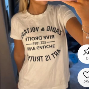 Vit t-shirt med svart tryck från Zadig & Voltaire - Vit t-shirt från Zadig & Voltaire med svart texttryck framtill. Klassisk rund halsringning och korta ärmar. Perfekt för en avslappnad och trendig stil. HAR DEFEKTER SE BILD!