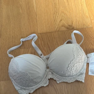Vit spets-bh med vadderade kupor - Säljer en vit bh med vadderade kupor och fina spetsdetaljer längs kanterna och under bysten. Axelbanden är justerbara och bh:n har en klassisk bygel för extra stöd. Perfekt för dig som vill ha både komfort och en feminin look.