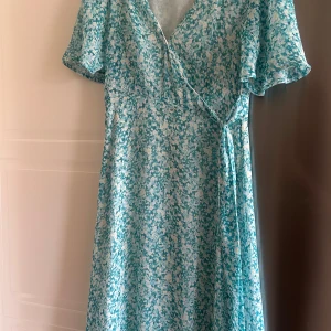 Sommarklänning  - Säljer en söt blå och vit blommig omlottklänning med korta ärmar och v-ringning. Klänningen har ett knyt i midjan och ett luftigt, somrigt tyg som passar perfekt till varma dagar.