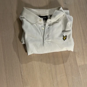 Vit pikétröja från Lyle & Scott - Säljer en klassisk vit pikétröja från Lyle & Scott med korta ärmar och gul broderad logga på bröstet. Tröjan har krage och knappar framtill. Perfekt för en stilren look.