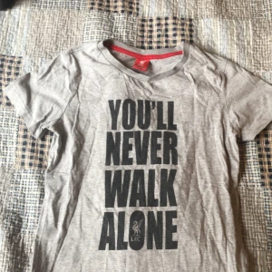 Grå t-shirt med Liverpool-tryck - Grå t-shirt med stort svart tryck på framsidan där det står 'YOU'LL NEVER WALK ALONE' och Liverpool FC:s logga längst ner. Rund halsringning och korta ärmar. Perfekt för dig som älskar fotboll och vill visa ditt stöd för Liverpool.