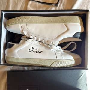 Vita sneakers från Saint Laurent - Säljer ett par stilrena vita sneakers från Saint Laurent med svart detalj på hälen och handskriven logga på sidan. Skorna har snörning och en beige platt sula. Perfekta för dig som gillar klassisk och clean design.