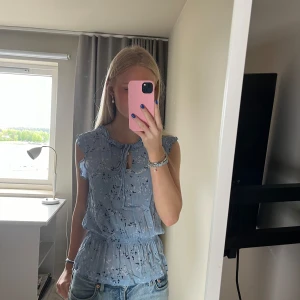 Ljusblå blommig blus med volang - Säljer en ljusblå blus med blommigt mönster och volangdetaljer. Blusen är ärmlös och har en liten knytning vid halsen samt resår i midjan för en snygg passform.☺️