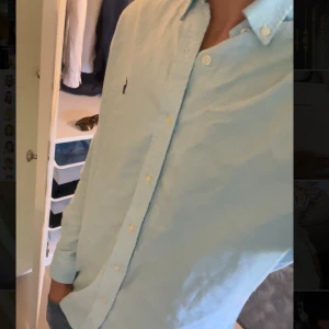 Ljusblå skjorta från Ralph Lauren - Säljer en klassisk ljusblå skjorta från Ralph Lauren med broderad logga på bröstet. Skjortan har lång ärm, knappar framtill och en stilren krage. Perfekt för en clean och stilren look.