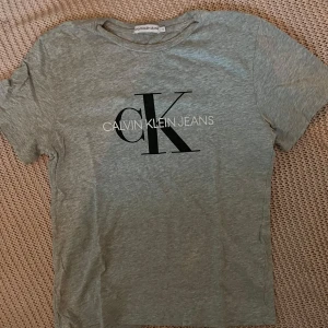 Grå t-shirt från Calvin Klein Jeans - Snygg grå t-shirt från Calvin Klein Jeans med stort svart CK-tryck och vit text på bröstet. Klassisk rund halsringning och korta ärmar. Perfekt till jeans eller shorts för en clean och stilren look.