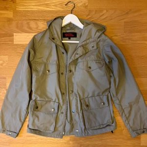 Grön jacka från Fjällräven G-1000 - Säljer en grön jacka från Fjällräven i G-1000-material. Jackan har huva, fyra fickor framtill med tryckknappar och dragkedja. Klassisk outdoor-look med diskret logga på ärmen och bröstficka.