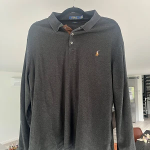 Svart långärmad pikétröja från Polo Ralph Lauren - Klassisk svart långärmad pikétröja från Polo Ralph Lauren i slim fit. Tröjan har krage, knappar vid halsen och den ikoniska broderade loggan på bröstet. Perfekt för en stilren och avslappnad look. Storlek XL