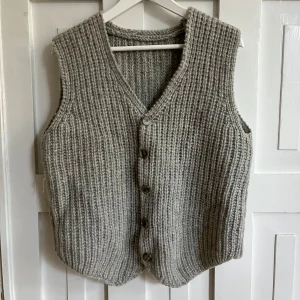Grå handstickad väst med knappar - Mysig grå handstickad väst med v-ringning och knäppning framtill. Västen har en grov ribbad struktur och är perfekt för lager-på-lager.