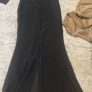 Svart glittrig långklänning med slits - Säljer en svart långklänning med glittrigt tyg och v-ringning. Klänningen har lång ärm och en snygg slits framtill. Perfekt för festliga tillfällen och har en elegant passform. Aldrig använt. 