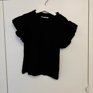 Svart topp med volangärmar från Gina Tricot - Säljer en svart topp från Gina Tricot med korta volangärmar. Toppen har en rund halsringning och är tillverkad i mjukt material. Aldrig använd och har lappen kvar.