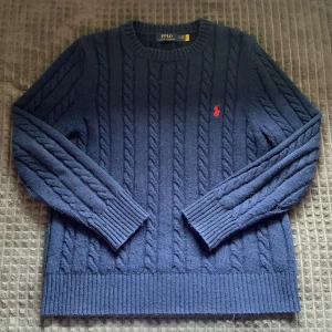Marinblå kabelstickad tröja från Polo Ralph Lauren - Säljer en marinblå kabelstickad tröja från Polo Ralph Lauren med rund halsringning och röd broderad logga på bröstet. Tröjan har långa ärmar och breda ribbade muddar vid ärmslut och nederkant. Perfekt för en klassisk och stilren look. Säljer den då den blivit för liten på mig. Den är inte använd många gånger så den är i mycket fint skick. 