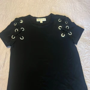 Michael Kors T-shirt  - Säljer en svart t-shirt från Michael Kors med coola snörningsdetaljer och metallöljetter på axlarna. T-shirten har korta ärmar och är gjord i mjukt material. Perfekt för dig som vill ha en stilren men ändå unik topp.