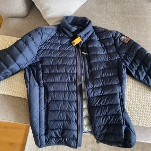 Mörkblå lättviktsjacka från Parajumpers - Säljer en mörkblå quiltad lättviktsjacka från Parajumpers med dragkedja och hög krage. Jackan har ett emblem på ena ärmen och snygga detaljer som gul rem vid halsen. Perfekt för kyliga dagar.