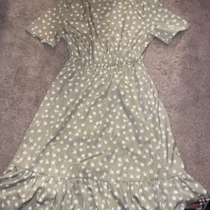 Ljusgrön blommig klänning med volang - Söt ljusgrön klänning med vitt blommönster och volang nertill. Klänningen har korta ärmar, resår i midjan och en luftig passform. Perfekt för vår och sommar!