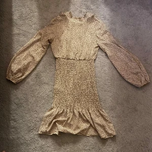 Beige klänning med brunt hjärtmönster och volang - Supersöt beige klänning med bruna hjärtan, långärmad och med volang nertill. Smockad midja för en bekväm passform och volangdetalj vid halsringningen. Perfekt för vår och sommar!