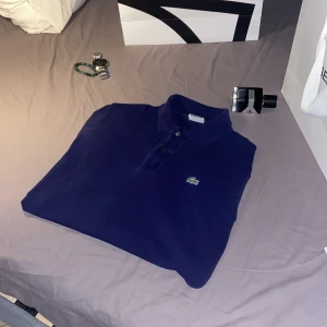 Mörkblå pikétröja från Lacoste - Säljer en klassisk mörkblå pikétröja från Lacoste med den ikoniska gröna krokodilloggan på bröstet. Tröjan har krage och knappar framtill, tillverkad i bomull för en bekväm känsla. Perfekt för en stilren och sportig look.