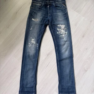 Blå Replay Grover jeans med slitningar - Säljer ett par blå Replay Grover jeans med snygga slitningar och tvättade detaljer. Klassisk femficksmodell med raka ben och normal passform. Perfekta för dig som gillar en avslappnad men stilren look.