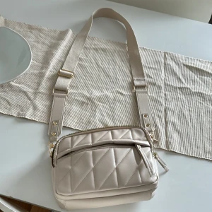 Beige quiltad axelväska med bred rem - Snygg beige axelväska med quiltad framsida och bred justerbar rem. Väskan har två fack med dragkedja i guld och är perfekt för att bära det viktigaste. Stilren och modern design som passar till många outfits.