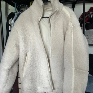 Beige teddyjacka med dragkedja - Mysig beige teddyjacka med hög krage och dragkedja framtill. Jackan har två sidofickor och är helfodrad. Perfekt för kyliga dagar och ger en trendig look med sin fluffiga struktur. Använd några få gånger