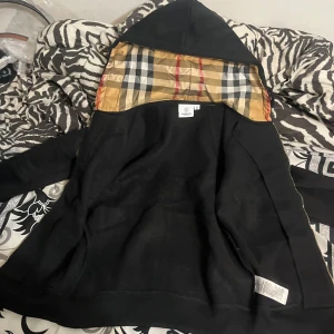 Svart hoodie med Burberry-rutig huva - Svart hoodie från Burberry med dragkedja och klassiskt Burberry-rutigt foder i huvan. Guldiga detaljer på dragkedjan och snörändarna. Perfekt för dig som gillar stilrena plagg med en twist. Den har aldrig använts innan