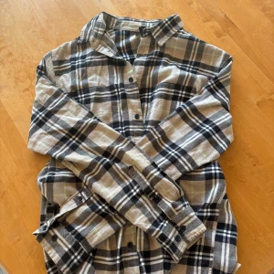 Beige och svart rutig skjorta från H&M - Säljer en klassisk rutig skjorta från H&M i beige, svart och vitt. storlek M. Skjortan har långa ärmar, knappar framtill och mjukt bomullsmaterial. Perfekt för en avslappnad stil och enkel att matcha med jeans eller chinos.