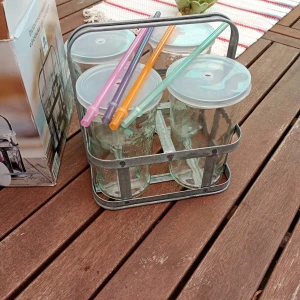 Fyra glas med sugrör och lock i metallställ från Victoria's Design House - Säljer ett set med fyra genomskinliga glas med lock och färgglada sugrör i ett praktiskt metallställ. Perfekt för sommarens drycker. Sugrören är i lila, orange, grön och rosa.