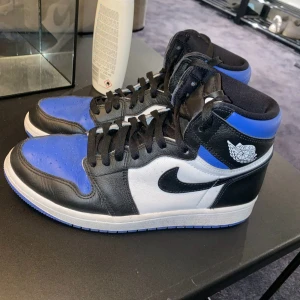 Nike Air Jordan 1 svart, vit och blå - Säljer ett par Nike Air Jordan 1’s med svart, vit och blå färgkombination. Skorna har klassisk hög modell, svart snörning och rund tå. Perfekta för dig som gillar streetwear och vill sticka ut med snygga sneakers.