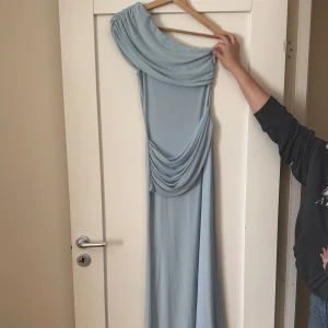 Ljusblå långklänning med drapering - Säljer en elegant ljusblå långklänning med unik drapering och en axel. Klänningen har en asymmetrisk design och är perfekt för speciella tillfällen. Materialet känns mjukt och följsamt mot kroppen.