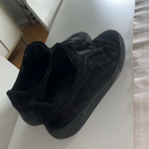 Arigato laceless mocha - Svarta slip-on sneakers från ARIGATO i mocka med platt sula och rund tå. Skorna har en stilren design utan snörning och en diskret logga på sidan. Perfekta för en clean och avslappnad look. Litet märke vid vänster fram pga av körning med crossmoped men inget som sticker ut för mycket. 