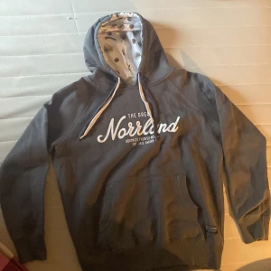 Grå Norrlands hoodie - Säljer en mörkgrå Norrlands hoodie med vit text 'The Great Norrland' på bröstet. Huvan är fodrad med vitt tyg och tryck av kottar och grenar. Klassisk känguruficka med grå och vita snören i huvan.