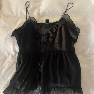 Svart volangblus från Monki - Säljer en svart, luftig blus från Monki med tunna axelband och volangdetaljer. Blusen är tillverkad i ett transparent, lätt material och har knytning framtill. tyvärr aldrig använd pga att den är för stor 💖