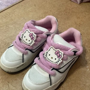 Hello Kitty sneakers med rosa detaljer - Säljer ett par vita sneakers med rosa detaljer och fluffiga rosa snören. Skorna har Hello Kitty-motiv både på plösen och innersulan. Perfekta för dig som älskar söta och unika skor! OBS! SKORNA ÄR REDAN TVÄTTADE (bilderna är gamla och skickar gärna nya bilder privat!)  använda ca 2 ggr.