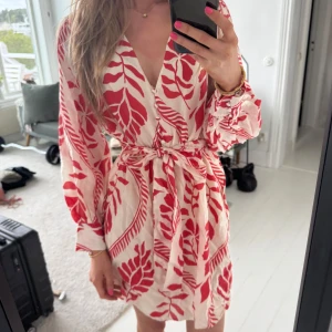 Vit och röd omlottklänning från H&M - Säljer en vit kortklänning från H&M med rött bladmönster. Klänningen har lång ärm, v-ringning, knappar framtill och knytskärp i midjan som går att ta av! Samt resor i midjan under bandet! Perfekt för sommaren!🥰 Använd 1 gång