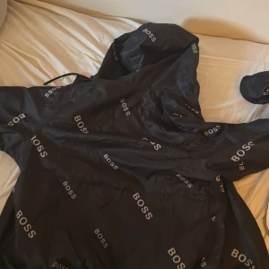 Svart vindjacka från BOSS - Snygg svart vindjacka från BOSS med logga tryckt över hela jackan. Jackan har huva och dragkedja framtill samt vita detaljer. Perfekt för dig som vill ha en stilren och sportig look.En helt ny kostar 5 tusen