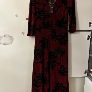 Röd långklänning med svart mönster från GIBS - Säljer en röd långklänning från GIBS med svart blommigt mönster. Klänningen har lång ärm och en figurnära passform upptill. Perfekt för dig som vill sticka ut med en unik och elegant look.