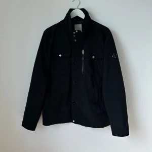 Svart Bailey jacka från J.Lindeberg - Stilren svart jacka från J.Lindeberg med hög krage, knappar och dragkedja framtill. Jackan har två bröstfickor med lock och knapp samt två sidofickor. Perfekt för dig som vill ha en klassisk look. Modell: Bailey Structured Poly -                                                Artnr: X239
