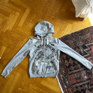 Grå hoodie med tryck från Pacha - Säljer en grå hoodie från Pacha med svart och färgglatt tryck på framsidan. Tröjan har dragkedja, huva och långa ärmar. Perfekt för en avslappnad stil och enkel att matcha med jeans eller leggings.