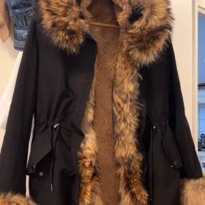 Svart parkajacka med fuskpäls - Säljer en svart parkajacka med brun fuskpäls runt huva, ärmslut och längs öppningen. Jackan har dragsko i midjan och stora fickor med lock. Insidan är fodrad med mjuk, brun pälsimitation för extra värme.