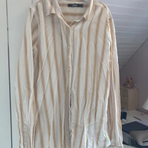 Beige och vit randig skjorta från Bik Bok - ✨Säljer en luftig skjorta från Bik Bok i beige och vitt med vertikala ränder. Skjortan har klassisk krage, knäppning framtill och långa ärmar. Perfekt att styla över ett linne eller knyta i midjan för en avslappnad look.✨