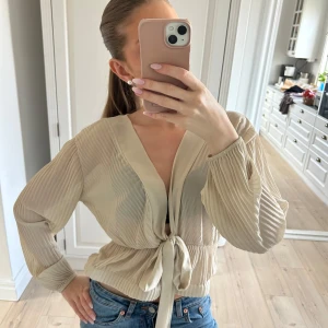 Beige blus med knyt framtill - Säljer en beige blus med långa, lätt transparenta ärmar och knyt framtill. Blusen har en v-ringad halsringning och plisserade detaljer på ärmarna💕