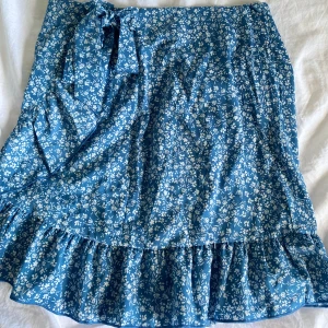 Blå blommig omlottkjol från SHEIN - Supersöt blå omlottkjol från SHEIN med vitt blommönster och volang längst ner. Kjolen har knytband i midjan och är perfekt för dig som gillar en feminin och somrig stil. I storlek S