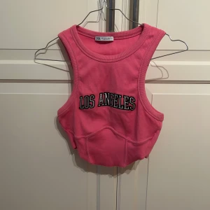 Rosa croppad linne från Zara - Säljer en rosa croppad linnetopp från Zara med texten 'Los Angeles' broderad framtill i strl s. Toppen är ribbad och ärmlös med korsett detalj vid bysten. 