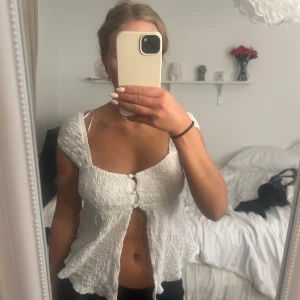 Vit blus med knappar från Gina Tricot - Supersöt vit blus från Gina Tricot med smockad struktur, breda axelband och tre klädda knappar framtill. Modellen är lätt utsvängd nertill och har en luftig känsla, perfekt för varma dagar. Passar dig som gillar en feminin och trendig stil. Använd fåtal gånger 