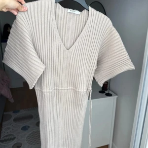 Beige ribbad klänning från NA-KD - Ribbstickad beige klänning från NA-KD med v-ringning, korta ärmar och knytband i midjan. Modellen är lång. Perfekt för dig som gillar stilrena och enkla plagg med snygga detaljer. 