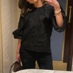 Svart blus med puffärmar - Säljer en svart blus med diskret mönster och snygga puffärmar. Blusen har rund halsringning och är tillverkad i ett tyg med struktur som ger en lyxig känsla. Perfekt för dig som vill ha något stilrent men ändå unikt.