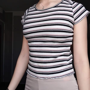 Randig ribbad topp från Lindex - Säljer en ribbad topp från Lindex med svarta, vita och ljusrosa ränder. T-shirten har rund halsringning och korta ärmar. Perfekt för dig som gillar en enkel men ändå snygg stil med lite retrovibbar.