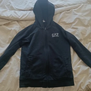 Svart EA7 hoodie med dragkedja - Svart hoodie från EA7 Emporio Armani med vit logga på bröstet. Tröjan har huva, hel dragkedja och två fickor framtill. Perfekt för dig som gillar stilrena och sportiga plagg. Passar dig som är ca 150 cm lång.