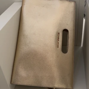 Guldig kuvertväska från Michael Kors - Stilren kuvertväska i guld från Michael Kors med elegant metallogo och praktiskt utskuret handtag. Väskan har en slät, lyxig yta och är perfekt för att lyfta din outfit med en touch av glamour.